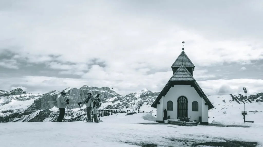Petite chapelle sur plateau enneigé, skieurs devant, crêtes des Dolomites