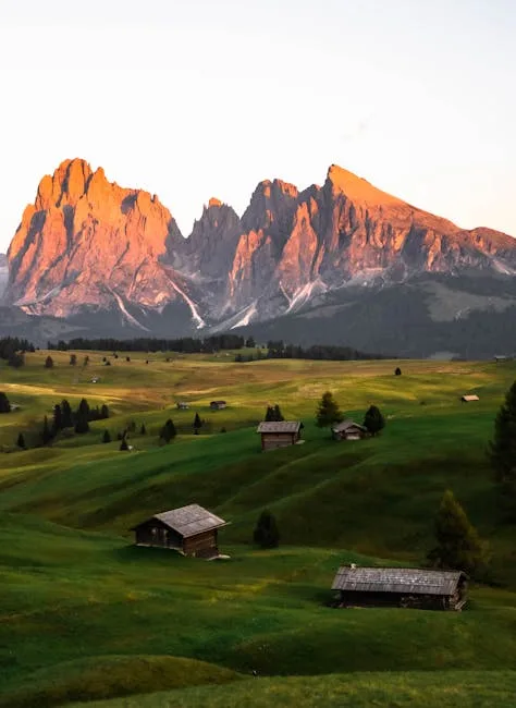 Cabanes dispersées sur un alpage verdoyant, sommets des Dolomites au coucher